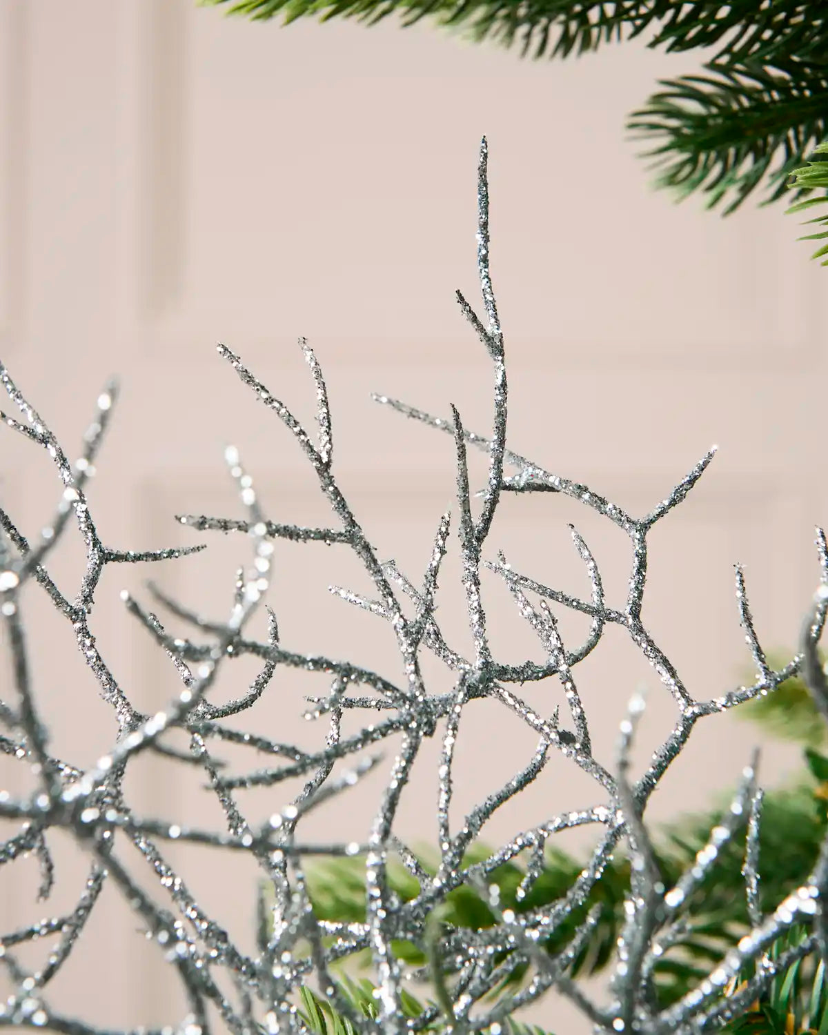 Silver glittery decorative branches with a sparkling finish, shown in close-up against a soft beige background with hints of green foliage.