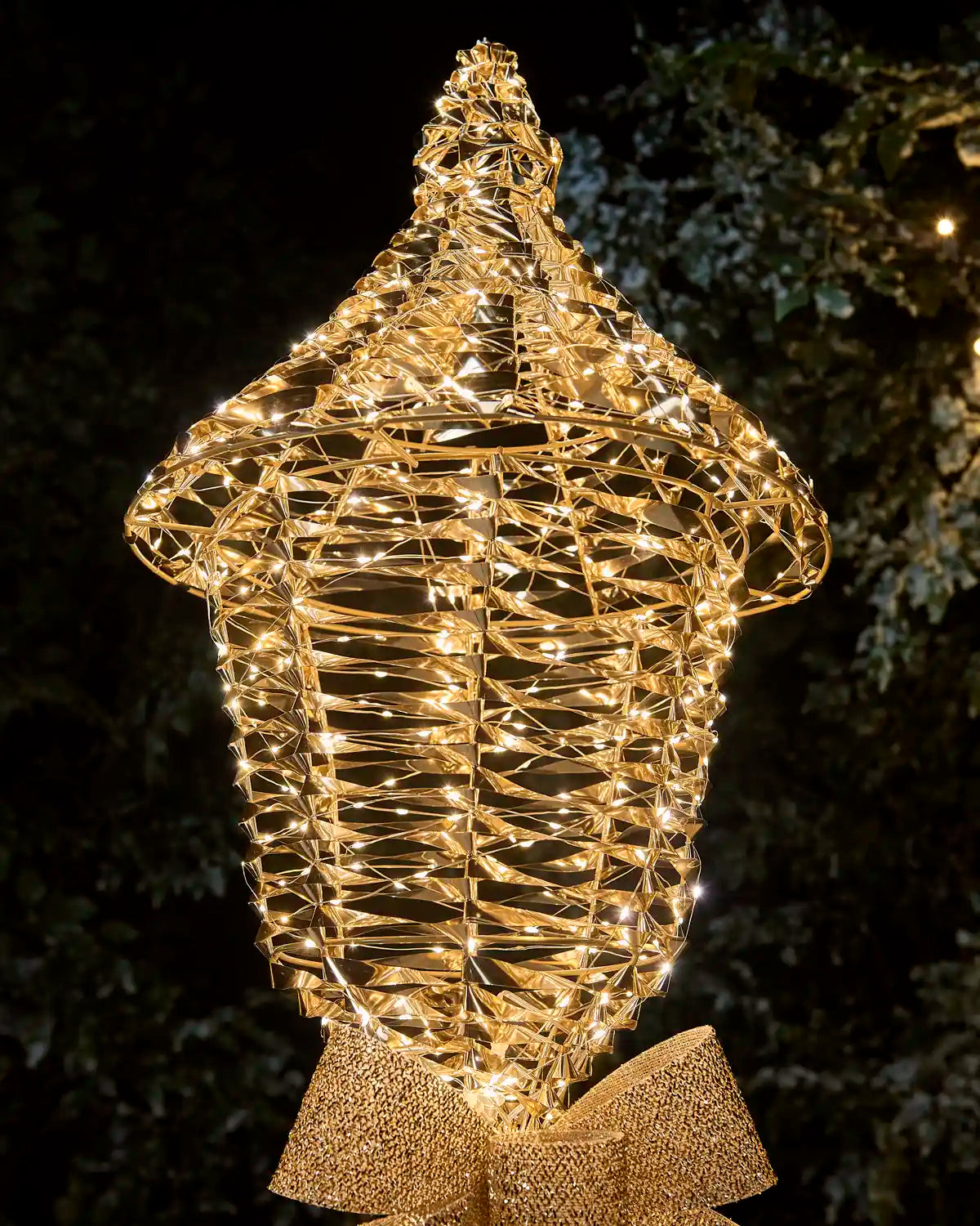 close up of a gold wire decorative light fixture shaped like a lantern or ornament, filled with warm white string lights, photographed outdoors against a dark night background with foliage.