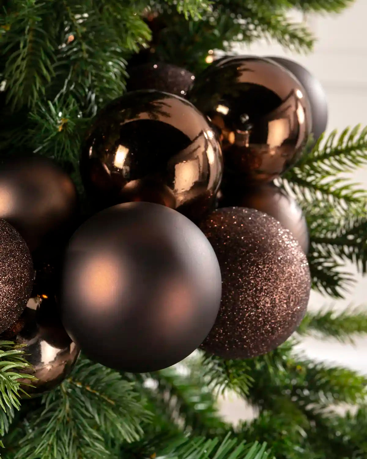 Close-up of espresso baubles showing shiny, matt and glitter finishes against green Christmas tree branches.
