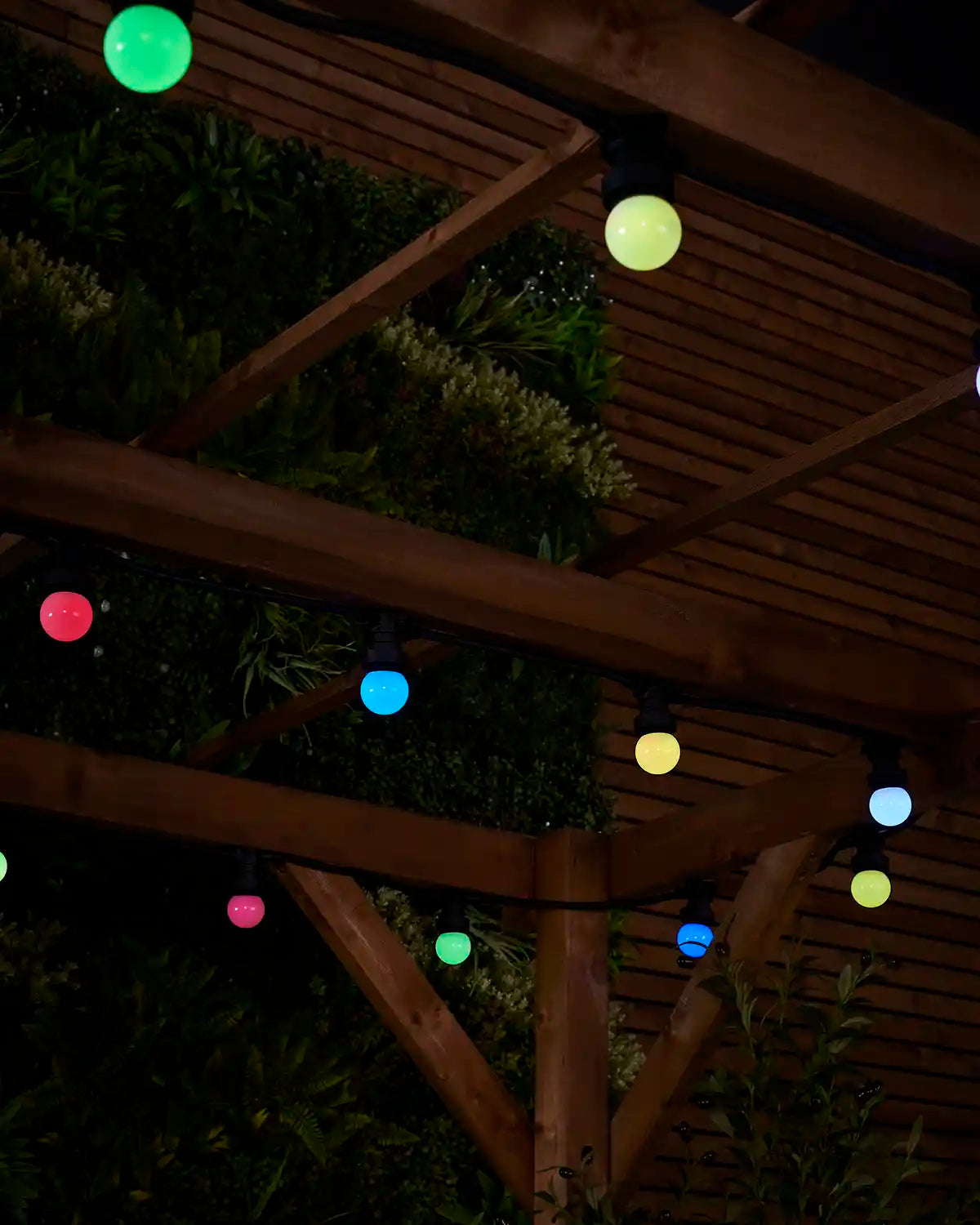 Close-up of colour-changing globe festoon bulbs hanging from a wooden pergola at night.