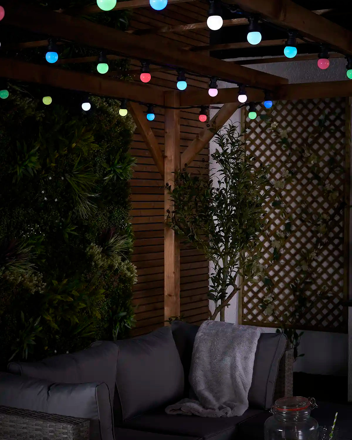 Colour-changing festoon lights shown across a patio pergola with garden plants and seating below.