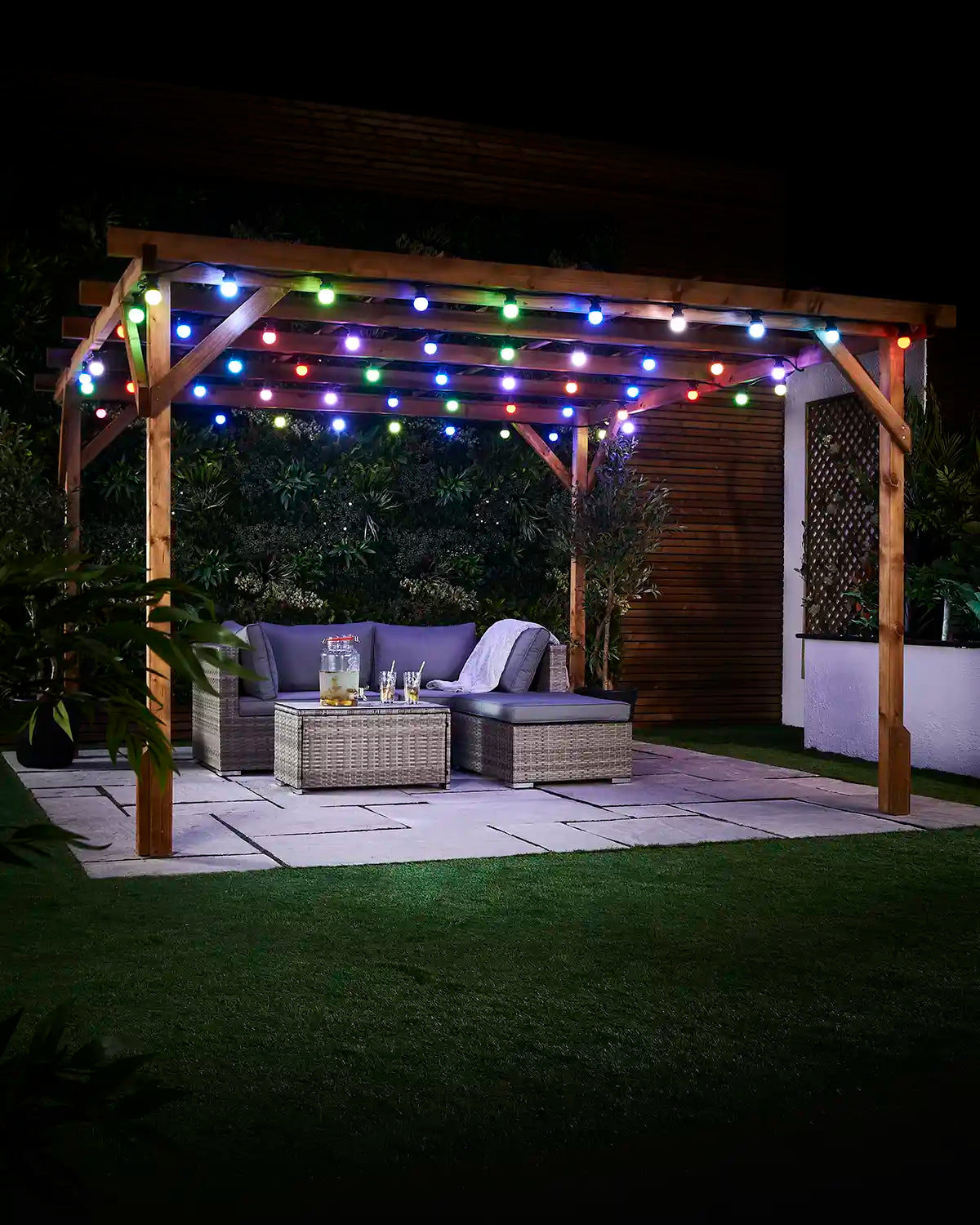 Colour-changing festoon bulbs displayed across an outdoor pergola above garden seating at night.