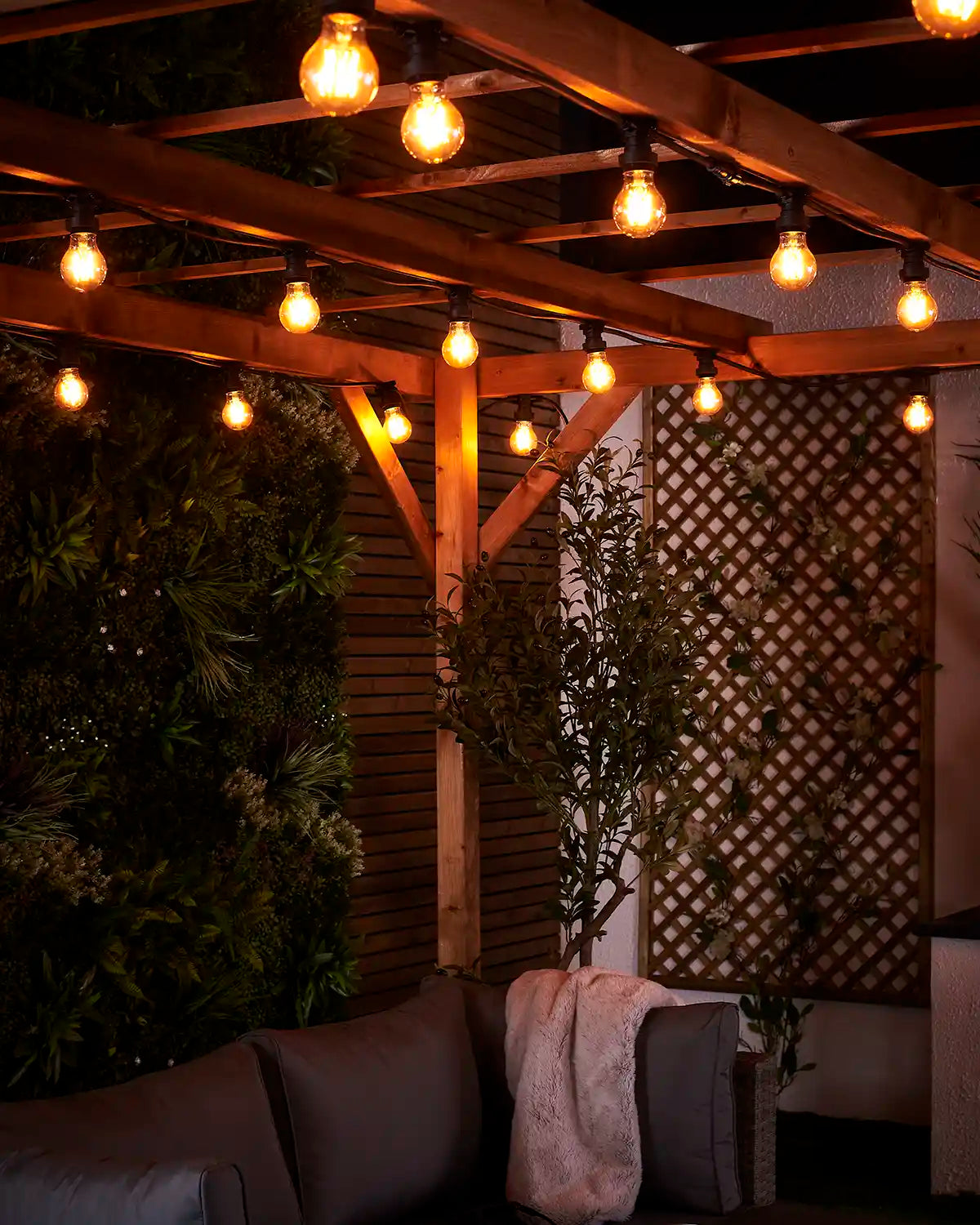 Warm white E27 festoon bulbs glowing above an outdoor sofa area beneath a wooden pergola.