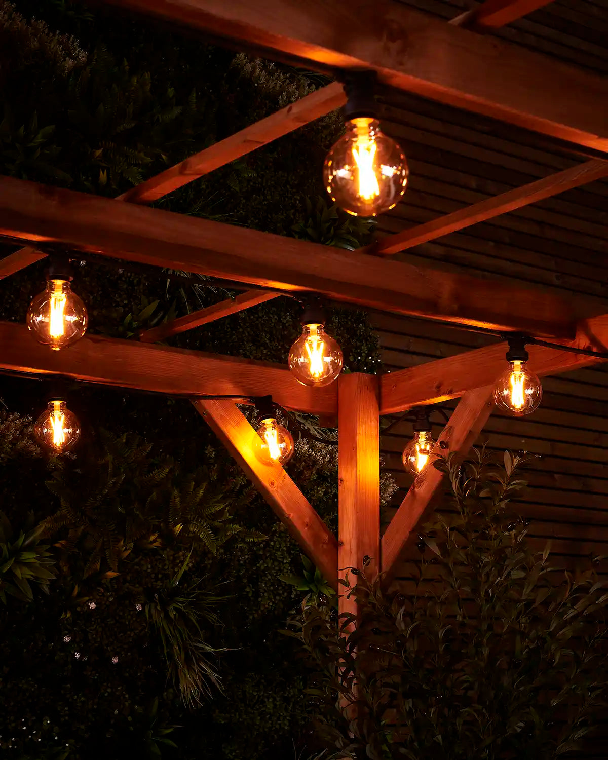 Close-up of warm white globe filament bulbs glowing under a wooden pergola with garden foliage behind.