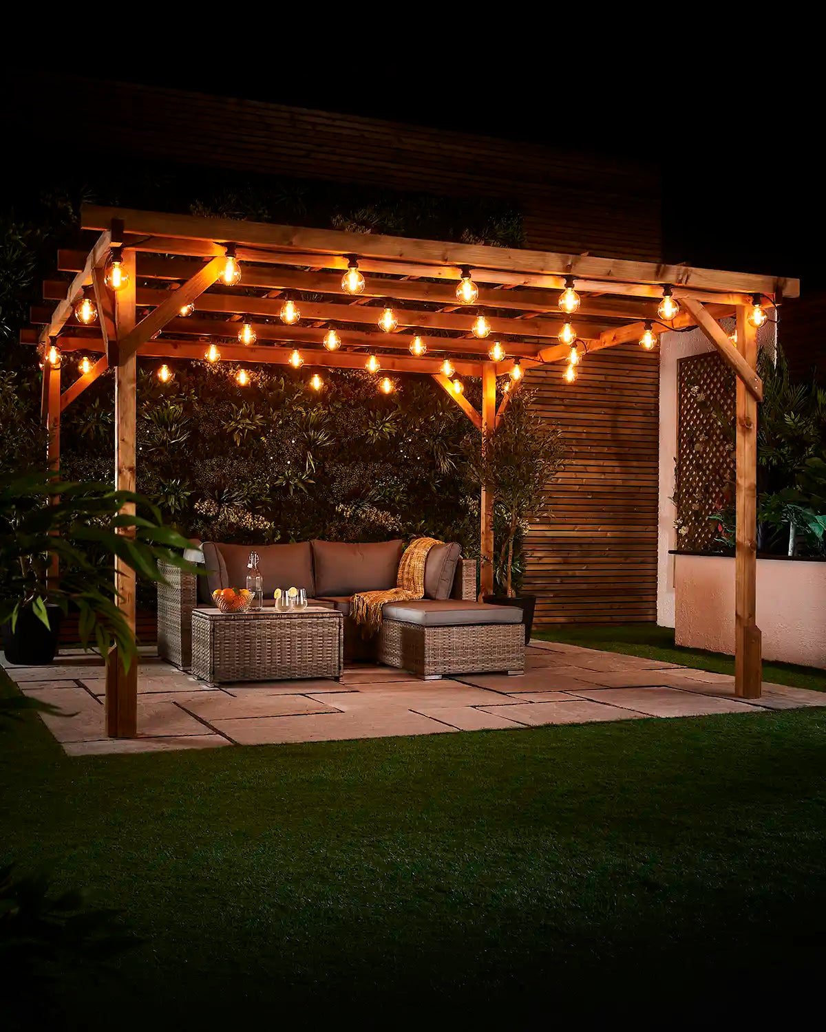 Warm white globe filament bulbs hanging across a wooden pergola above an outdoor seating area at night.