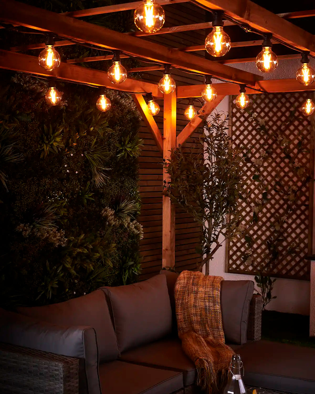 Warm white festoon globe bulbs lighting an outdoor sofa area beneath a wooden pergola at night.