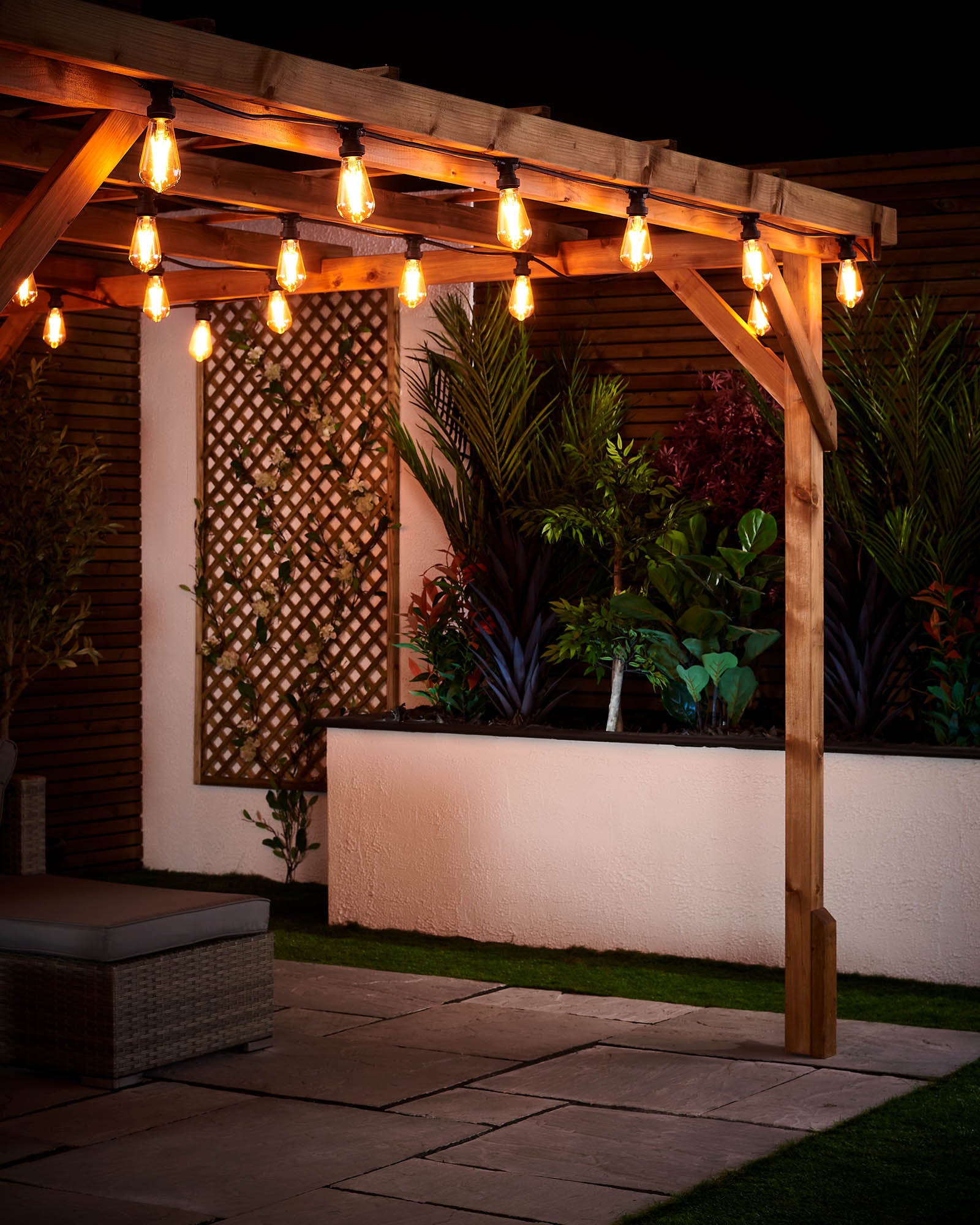 Warm white teardrop filament bulbs fitted along a wooden pergola in an outdoor patio setting with garden plants behind.