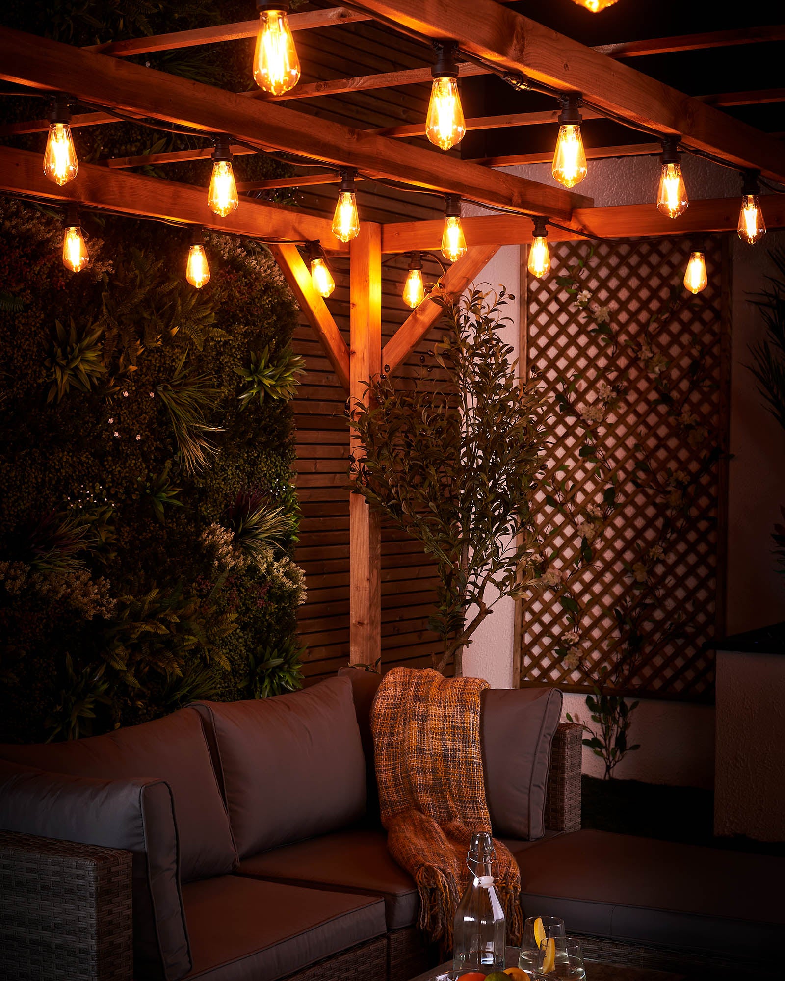 Warm white teardrop festoon bulbs lighting an outdoor sofa area beneath a wooden pergola at night.
