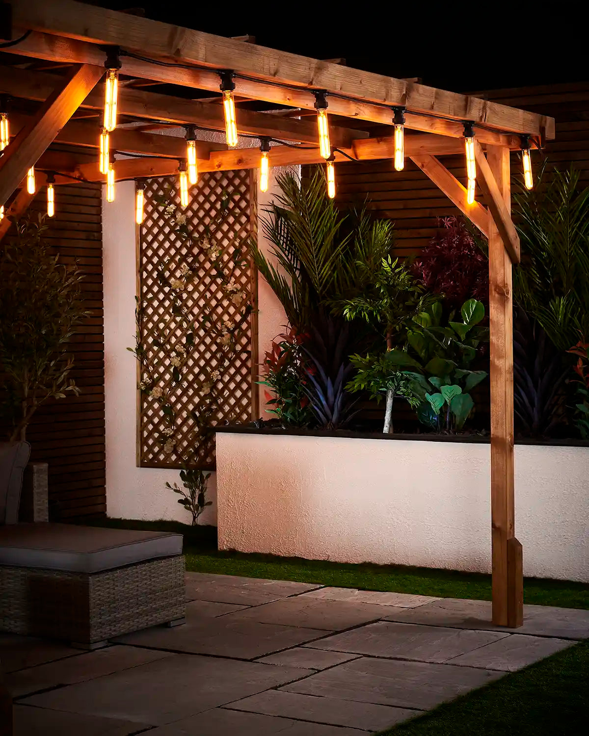 Warm white tubular filament bulbs fitted along a wooden pergola in an outdoor patio setting with garden plants behind.
