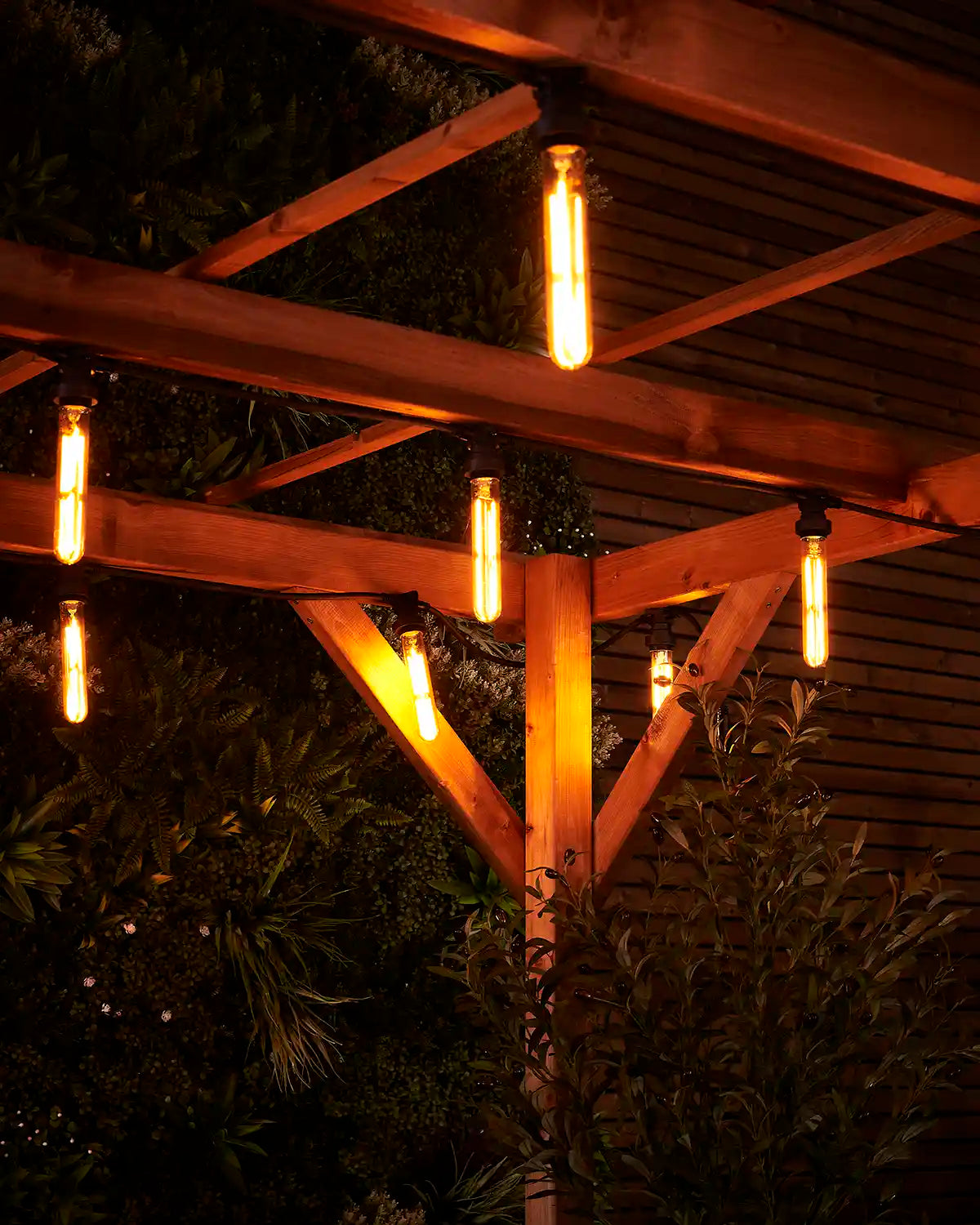 Close-up of warm white tubular filament bulbs glowing under a wooden pergola with garden foliage behind.