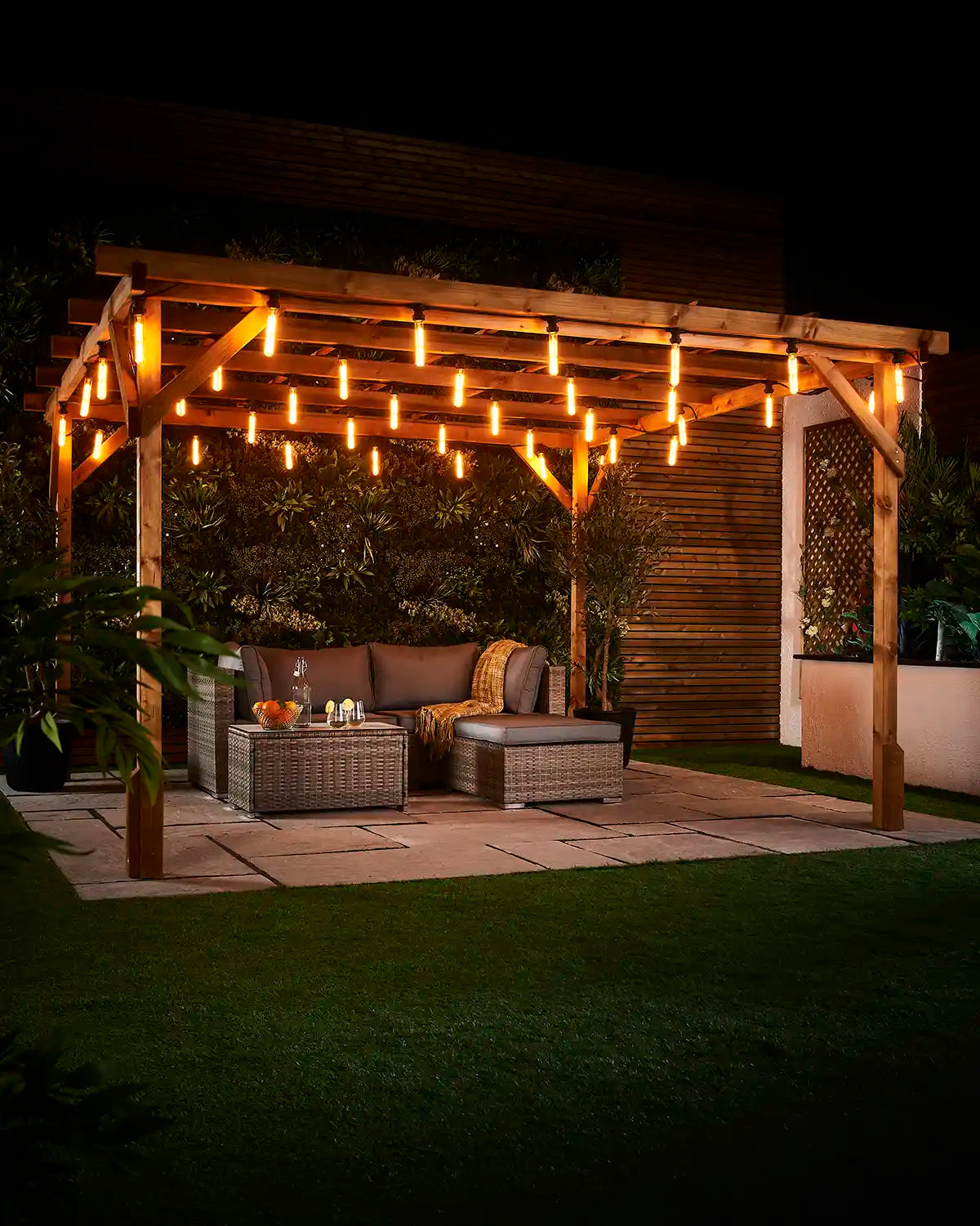 Warm white tubular filament bulbs hanging across a wooden pergola above an outdoor seating area at night.