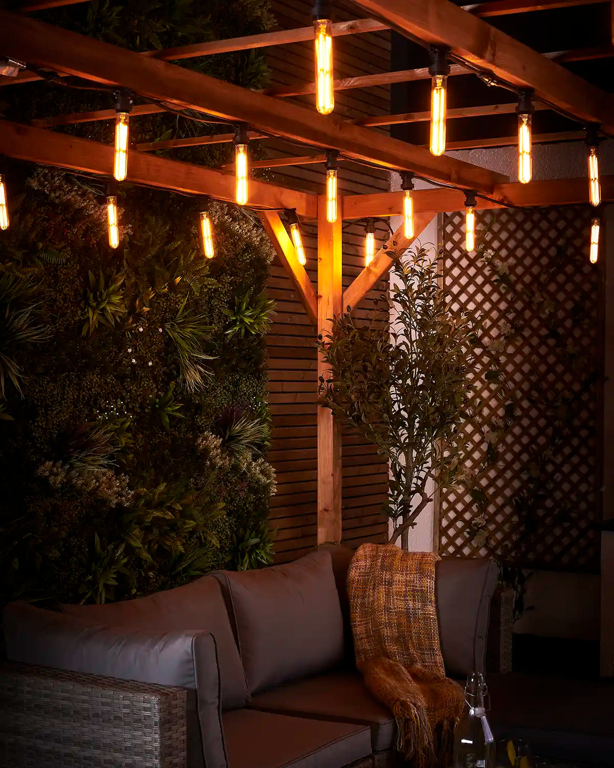 Warm white tubular festoon bulbs lighting an outdoor sofa area beneath a wooden pergola at night.