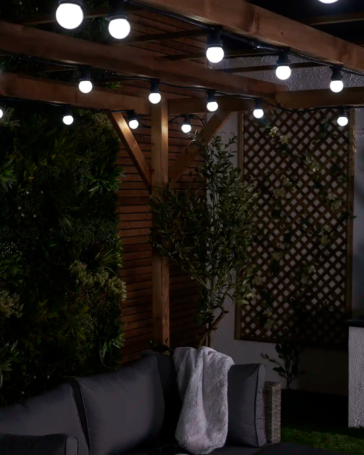 White festoon bulbs displayed across a wooden garden pergola with patio seating below.