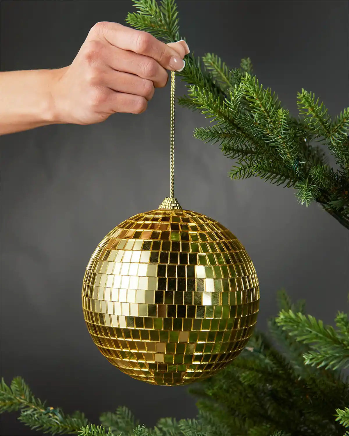 Hand holding a gold 15cm oversized disco ball bauble beside a Christmas tree.