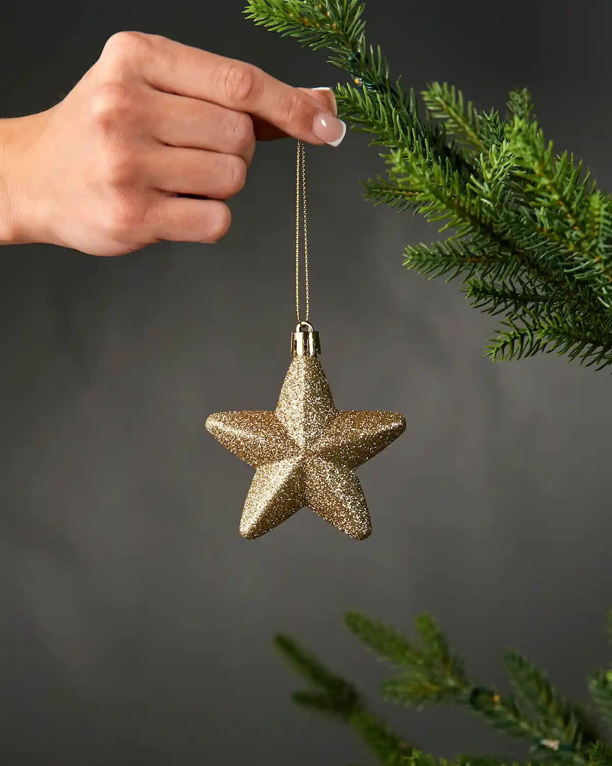 Hand holding a gold 7cm glitter star decoration beside a Christmas tree.