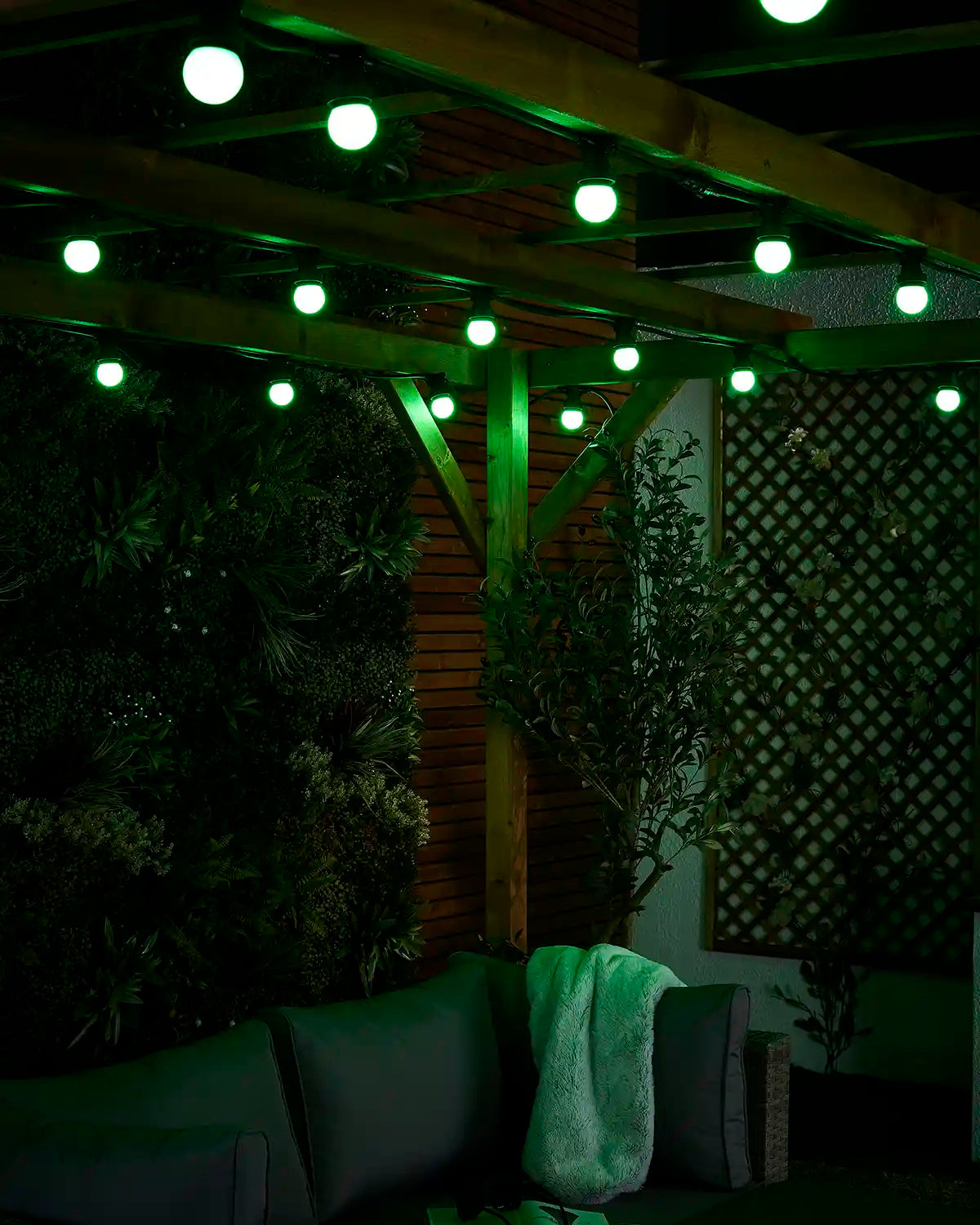 Close-up of green E27 festoon bulbs glowing beneath a wooden pergola with outdoor seating below.
