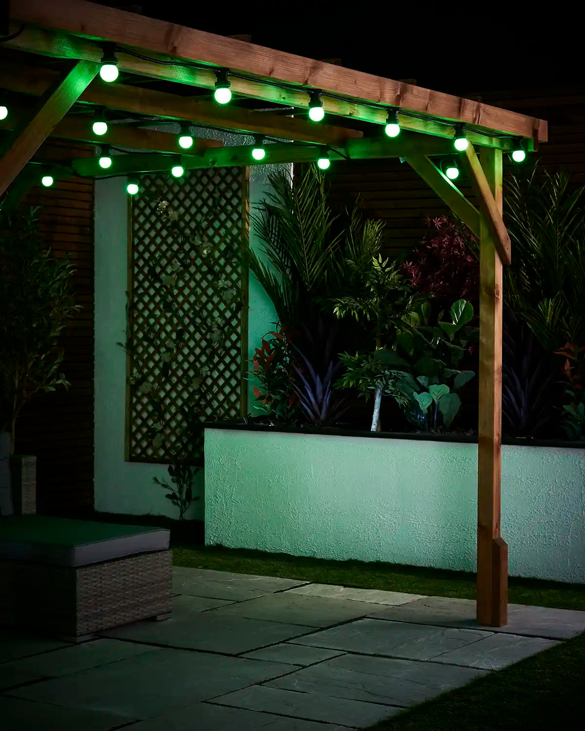Green E27 festoon bulbs hanging from a wooden pergola above a paved patio with a raised garden bed behind.