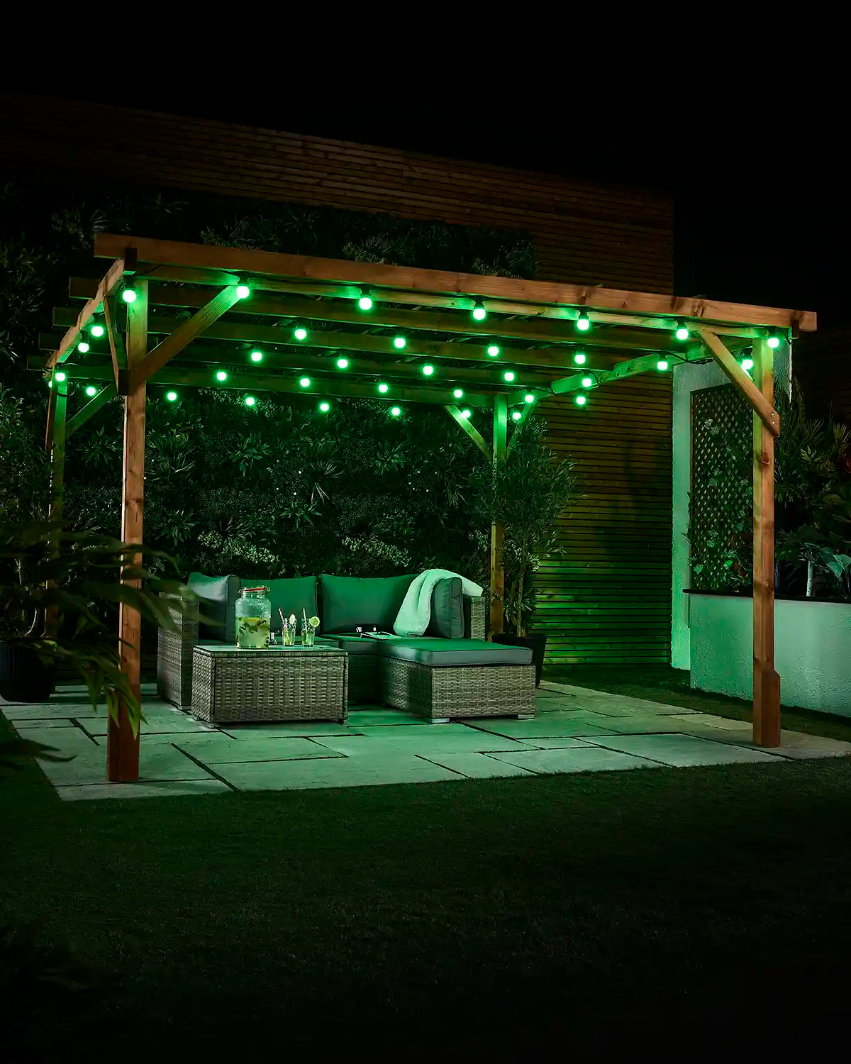 Green E27 festoon bulbs displayed across an outdoor wooden pergola at night with patio seating and garden plants.