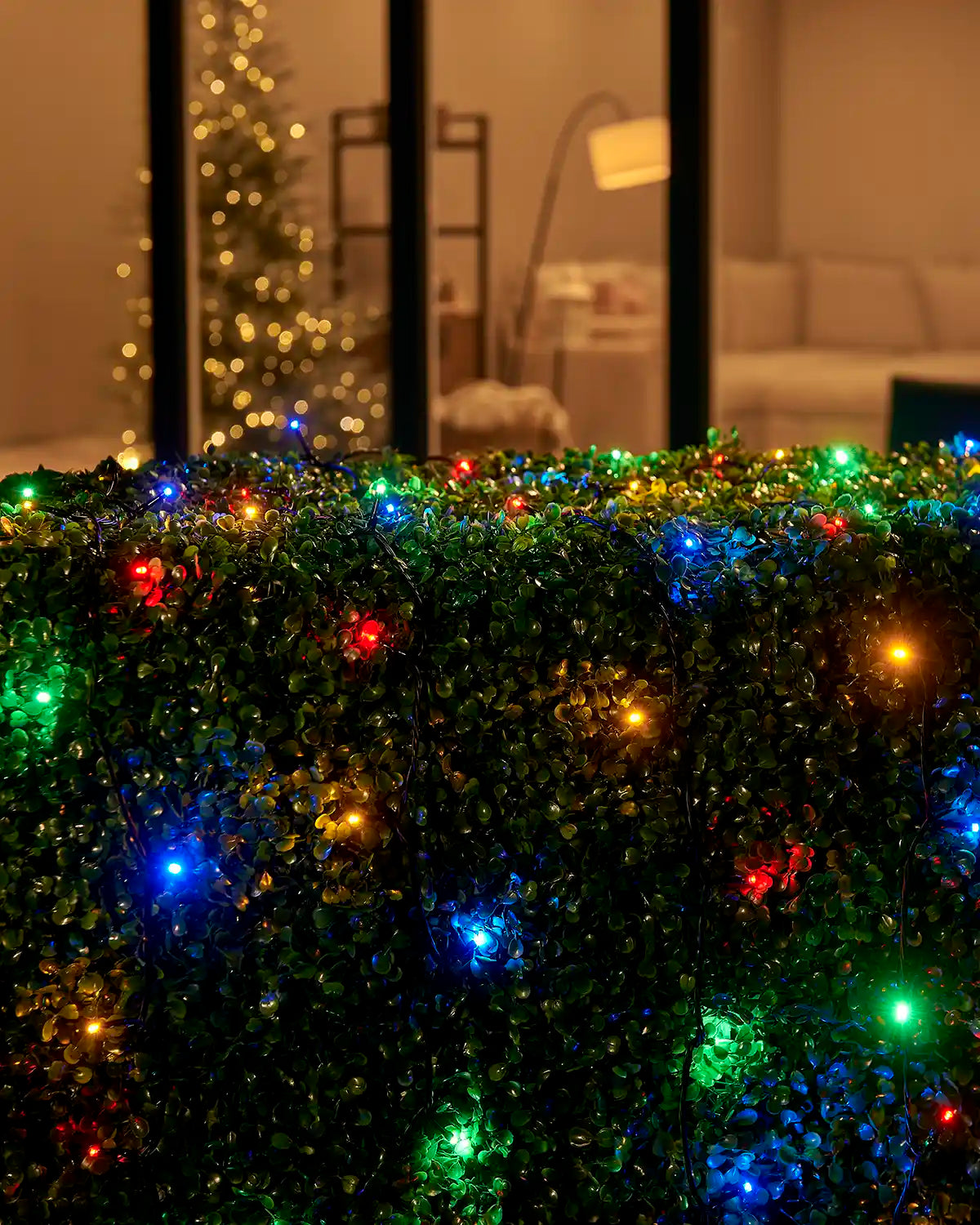 Multi colour Home Series string lights arranged across an outdoor hedge by patio doors.