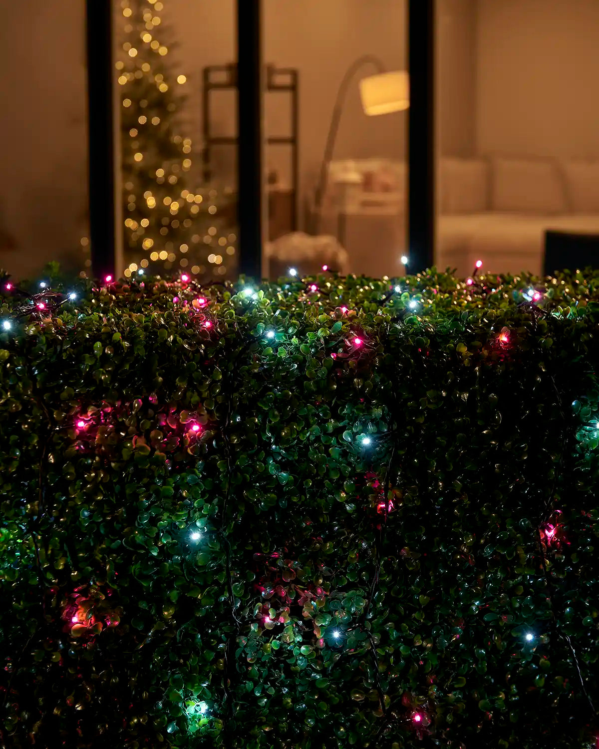 Pink and white Home Series string lights arranged across an outdoor hedge by patio doors.