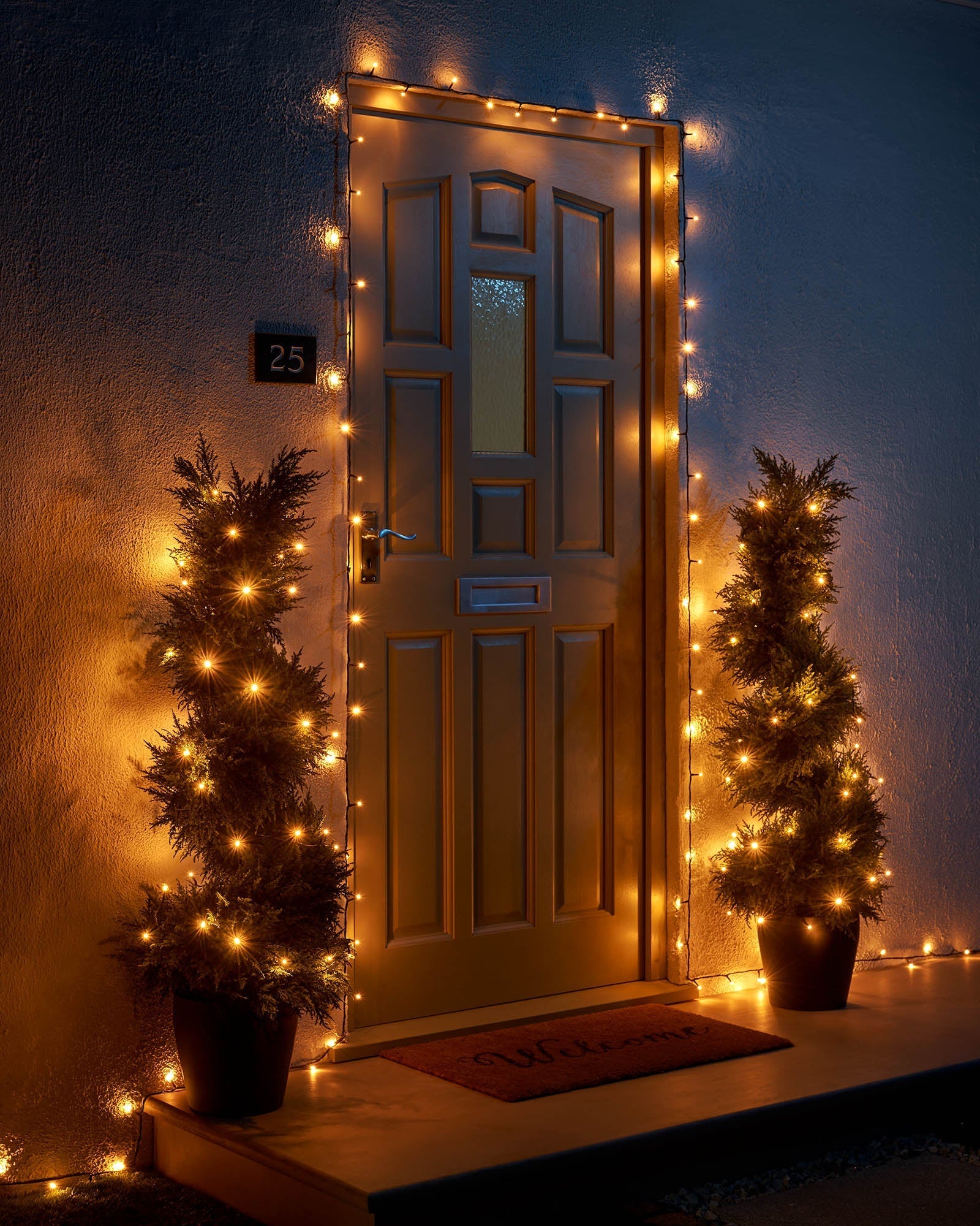 Warm white Home Series string lights framing a front door with two lit potted trees.