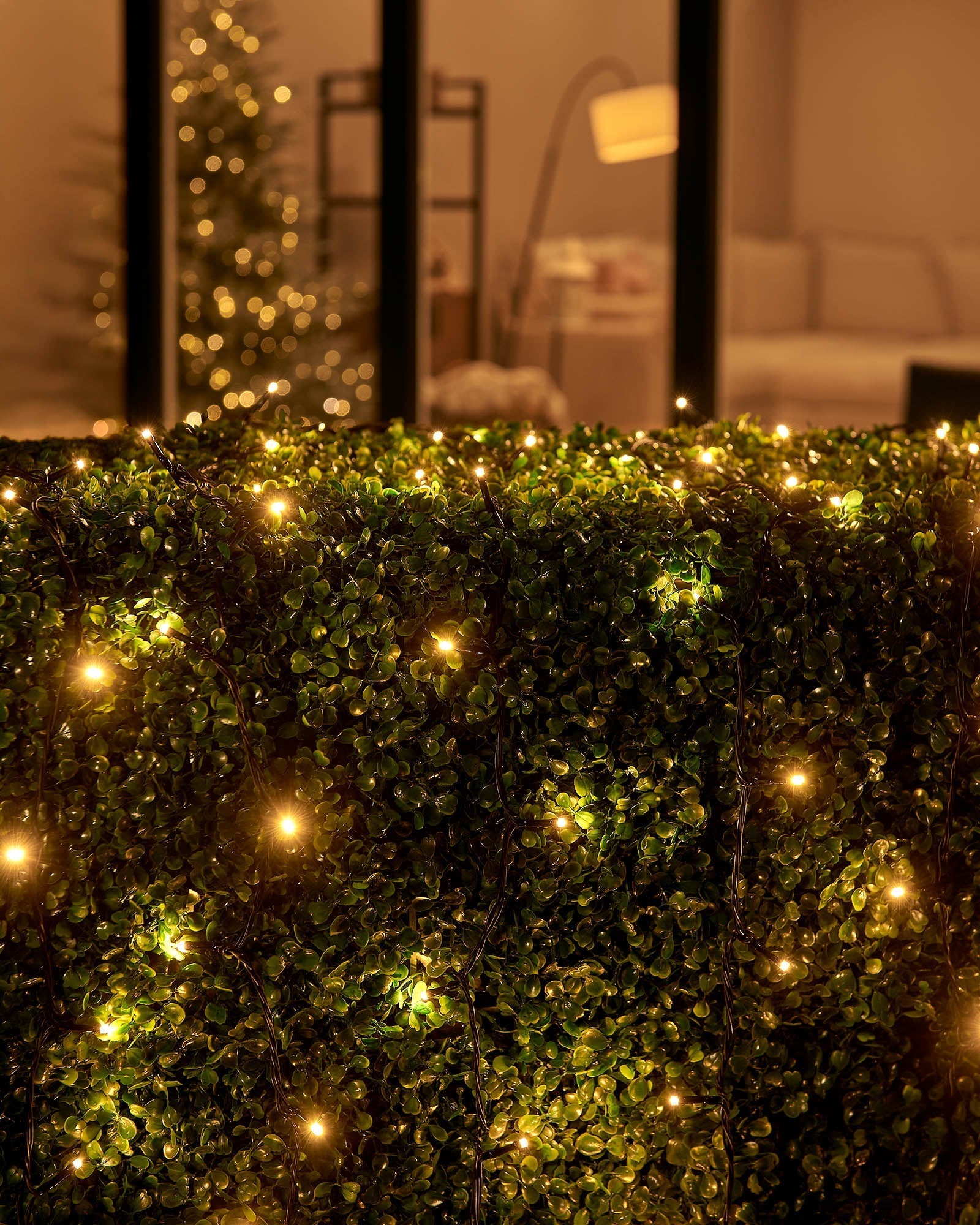 Warm white Home Series string lights arranged across an outdoor hedge by patio doors.