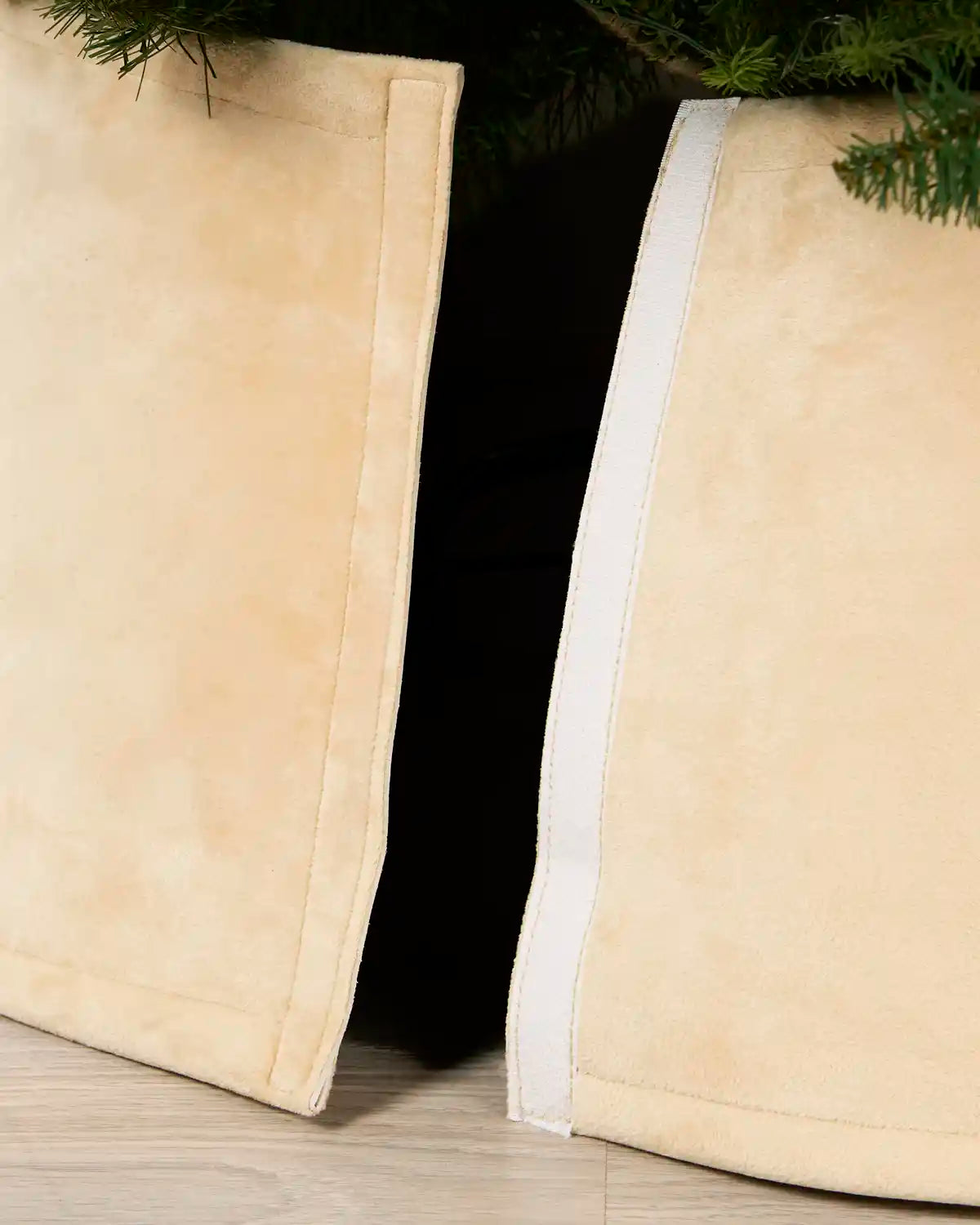 latte coloured Christmas tree skirt panels with white trim, shown partially folded to reveal a dark opening and green pine branches above.