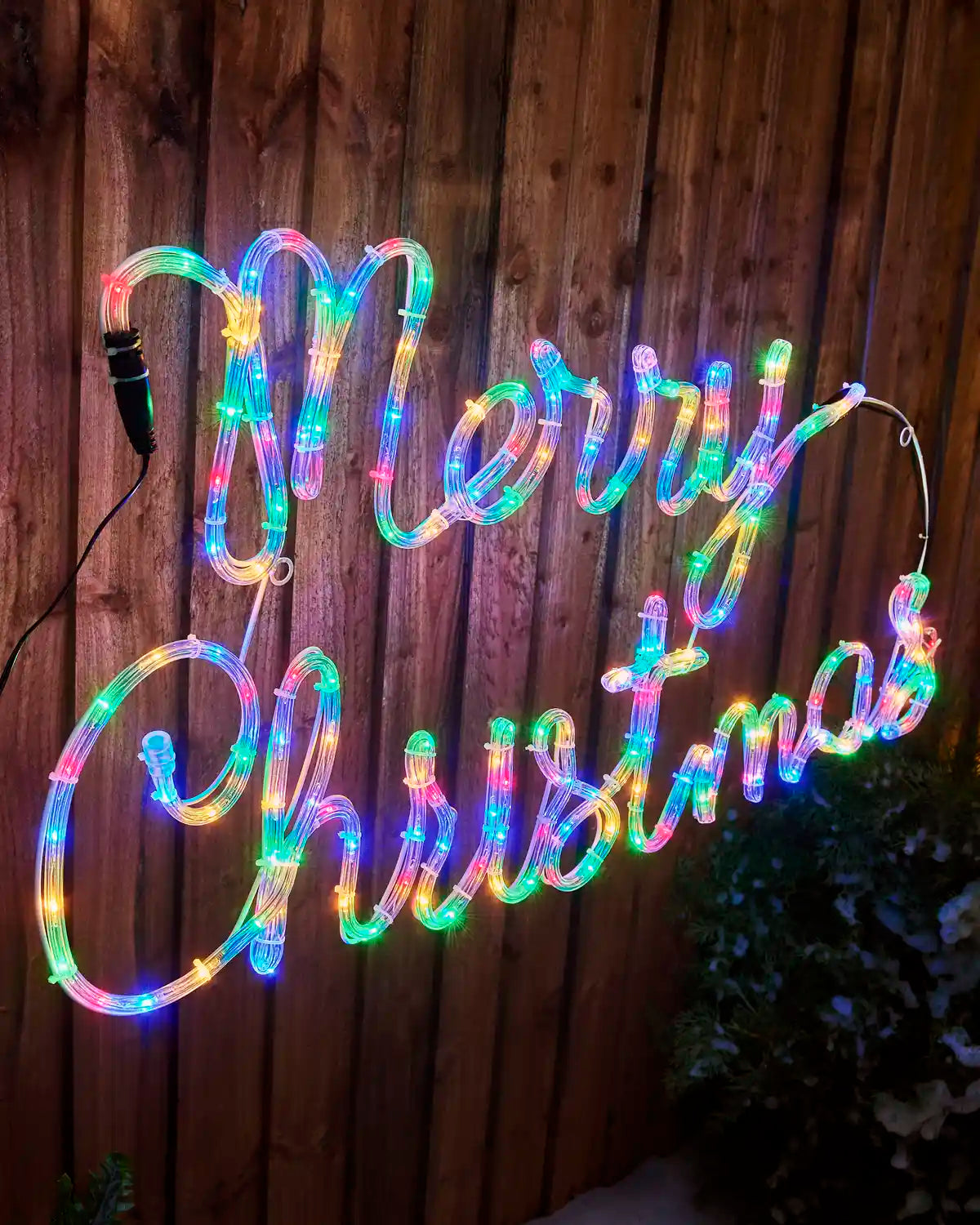 Angled view of the 100 cm Merry Christmas rope light display illuminated with multi-coloured LEDs on an outdoor wooden fence.