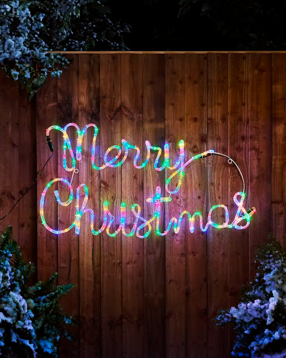 100 cm Merry Christmas rope light display with multi-coloured LEDs mounted on a wooden fence between snow-covered Christmas trees.