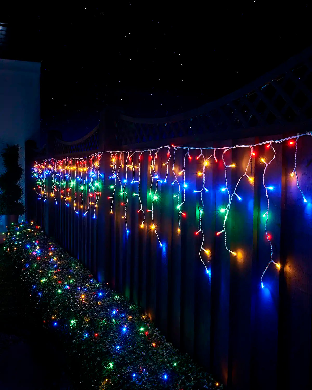 Multi-colour icicle lights with white cable displayed along a garden fence above a lit hedge at night.
