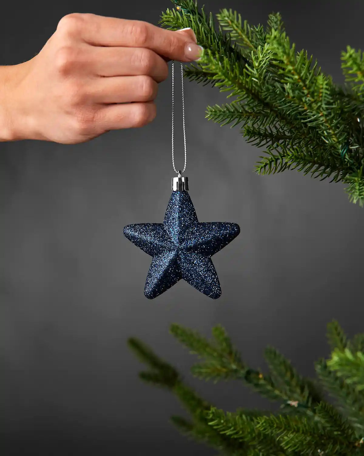 Hand holding a navy blue 7cm glitter star decoration beside a Christmas tree.