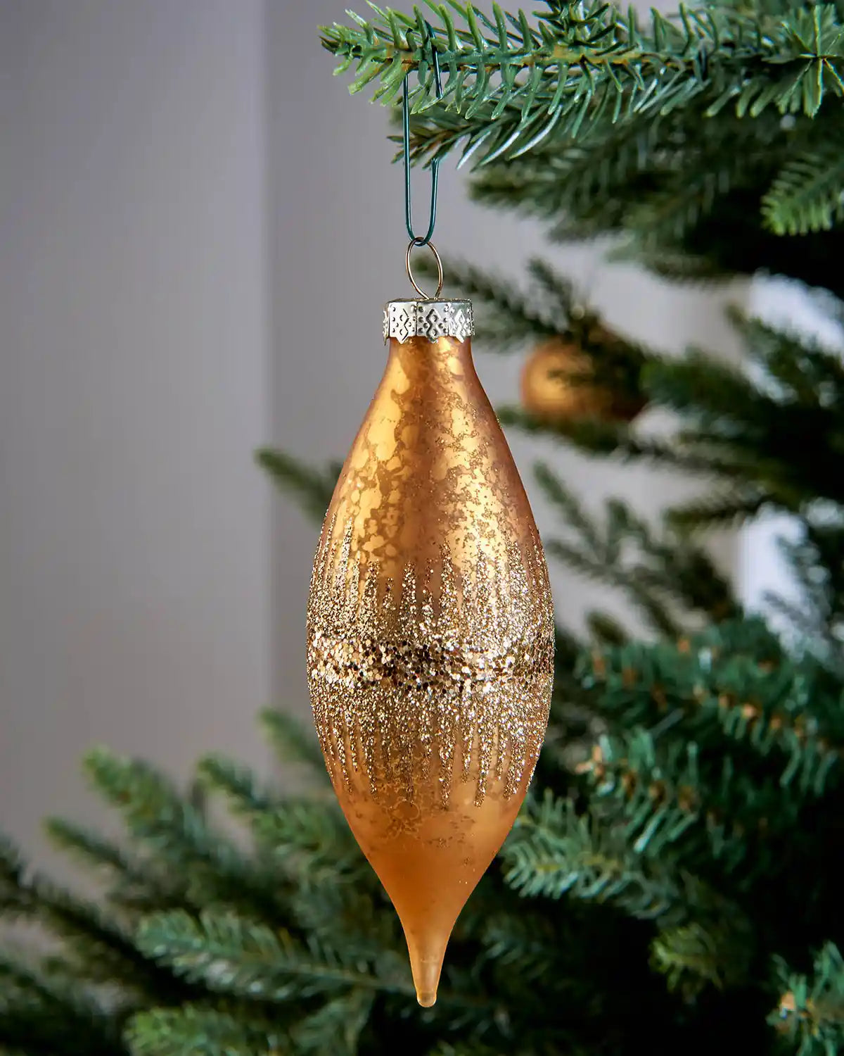 Gold teardrop Christmas ornament hanging from a green bauble hook on a tree branch