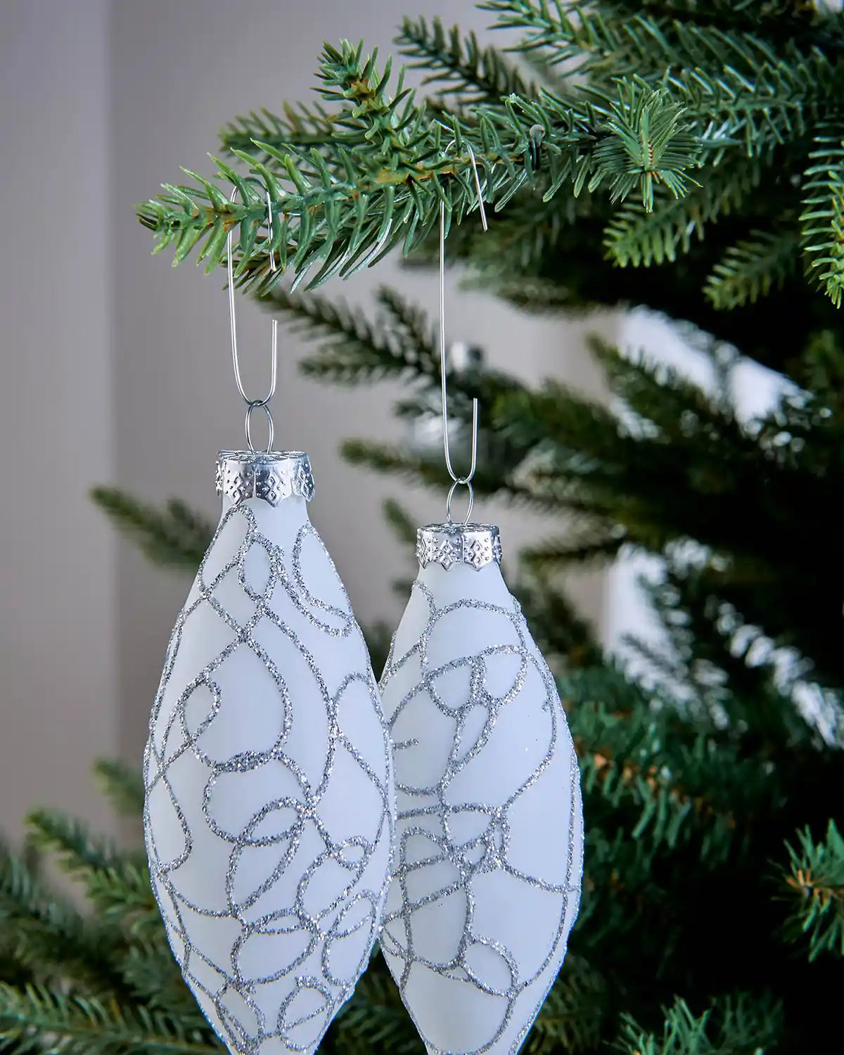 Two white Christmas ornaments hanging from silver bauble hooks on a tree branch