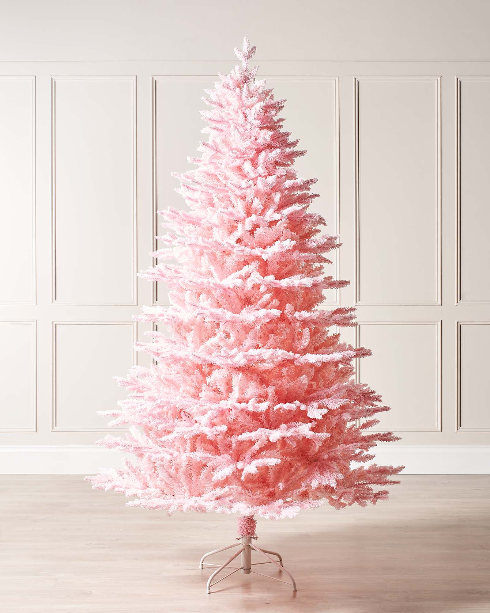 Pink snow flocked Christmas tree shown in full indoors against wall panelling, with a soft pink finish and metal tree stand.