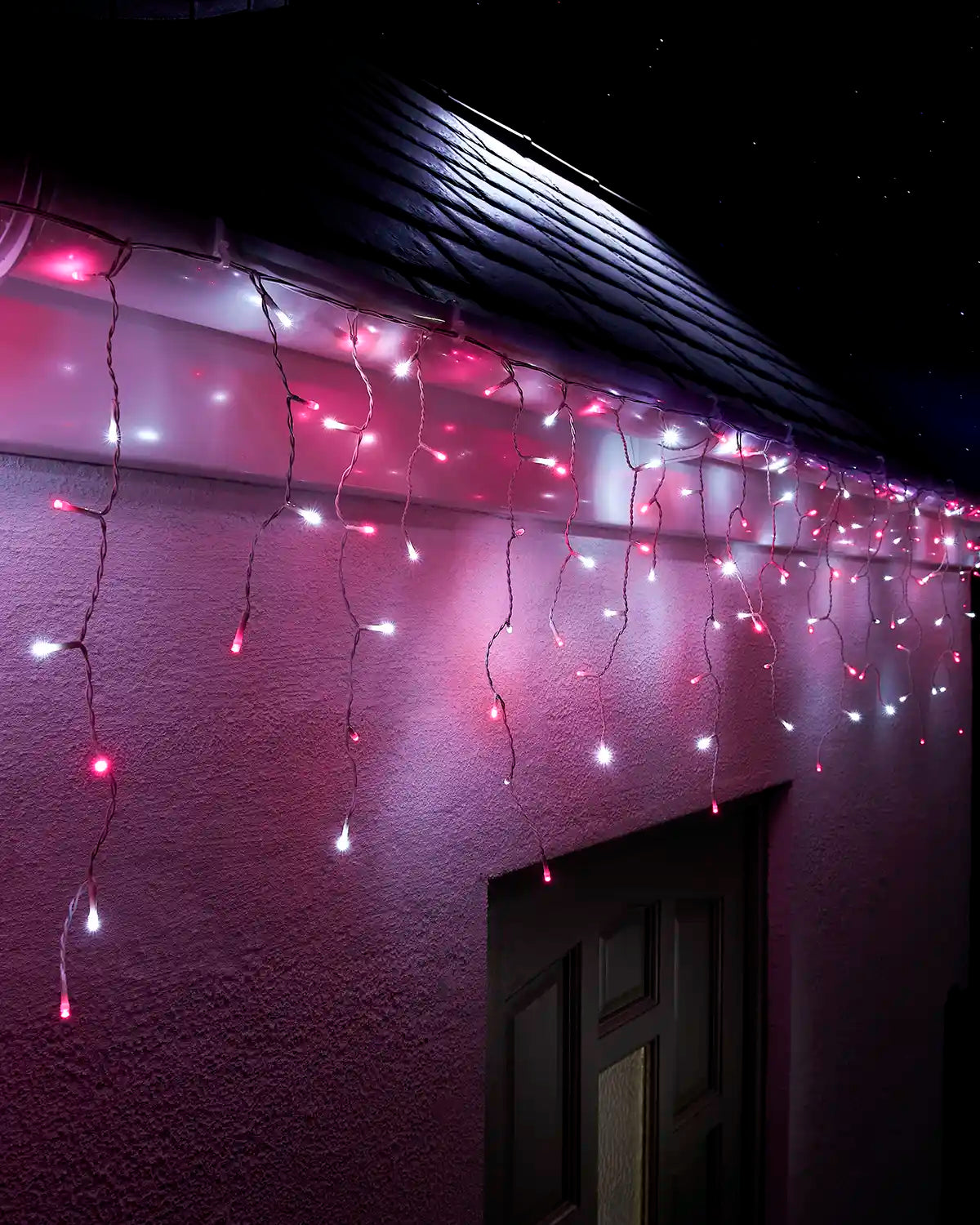 Pink and white icicle lights with white cable hanging along a roofline above an exterior wall and doorway at night.