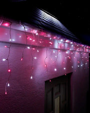 Pink and white icicle lights with white cable hanging along a roofline above an exterior wall and doorway at night.