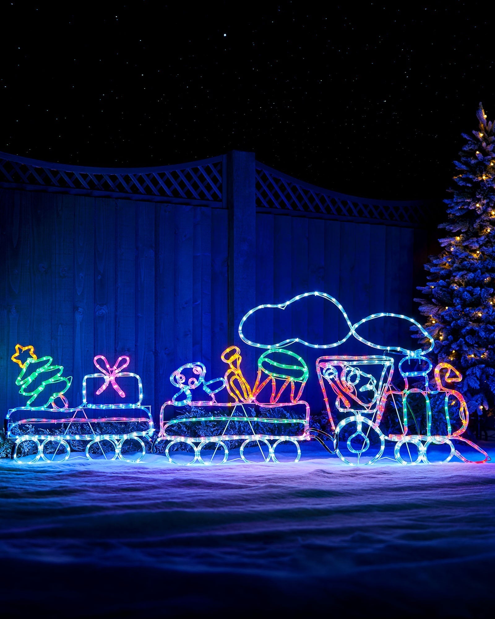 Multi-coloured animated steam train light displayed outdoors on snow beside a Christmas tree