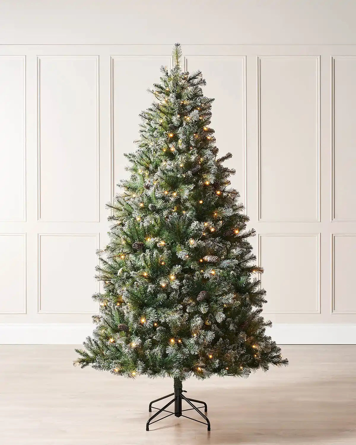 Full indoor view of a pre-lit Blue Brockton spruce Christmas tree with warm white lights, light frosted detailing, pinecones and a metal tree stand against wall panelling.