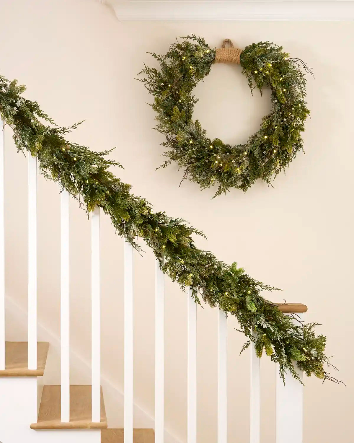 Pre-lit snow flocked wreath displayed above a matching illuminated garland on a staircase.