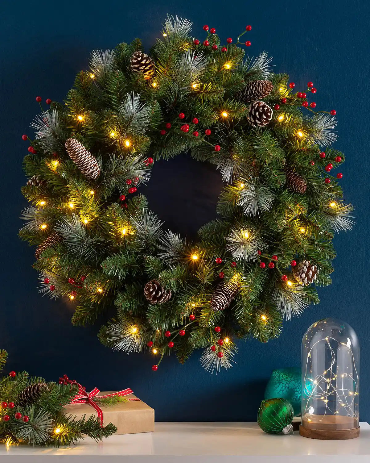 60 cm pre-lit frosted Christmas wreath with pinecones, red berries and warm white lights displayed on a wall