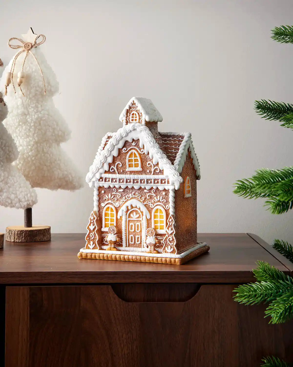 22cm pre-lit gingerbread house displayed on a wooden mantelpiece with soft festive tree decorations nearby.