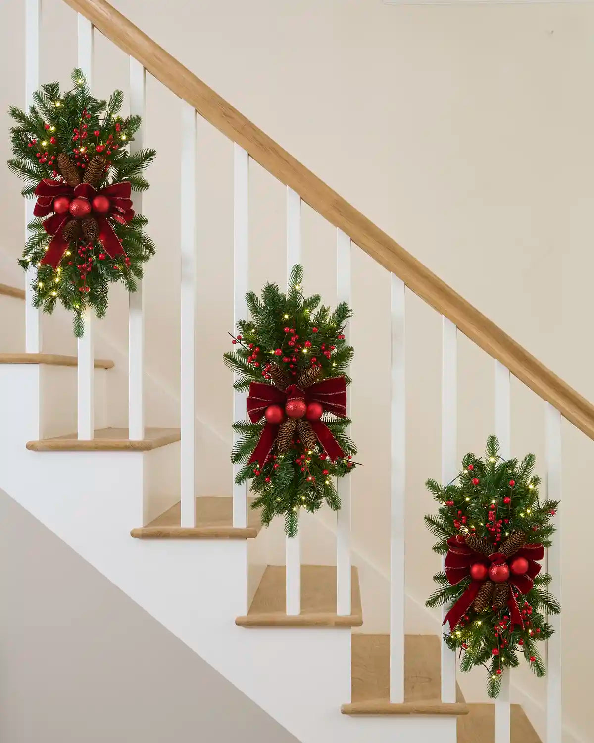 Three pre-lit red and burgundy garland swags displayed along a staircase banister.