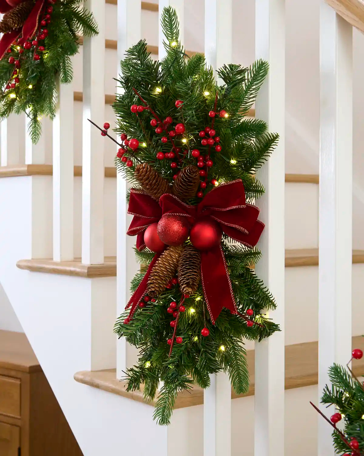 Red and burgundy pre-lit garland swag with warm white lights displayed on a staircase.