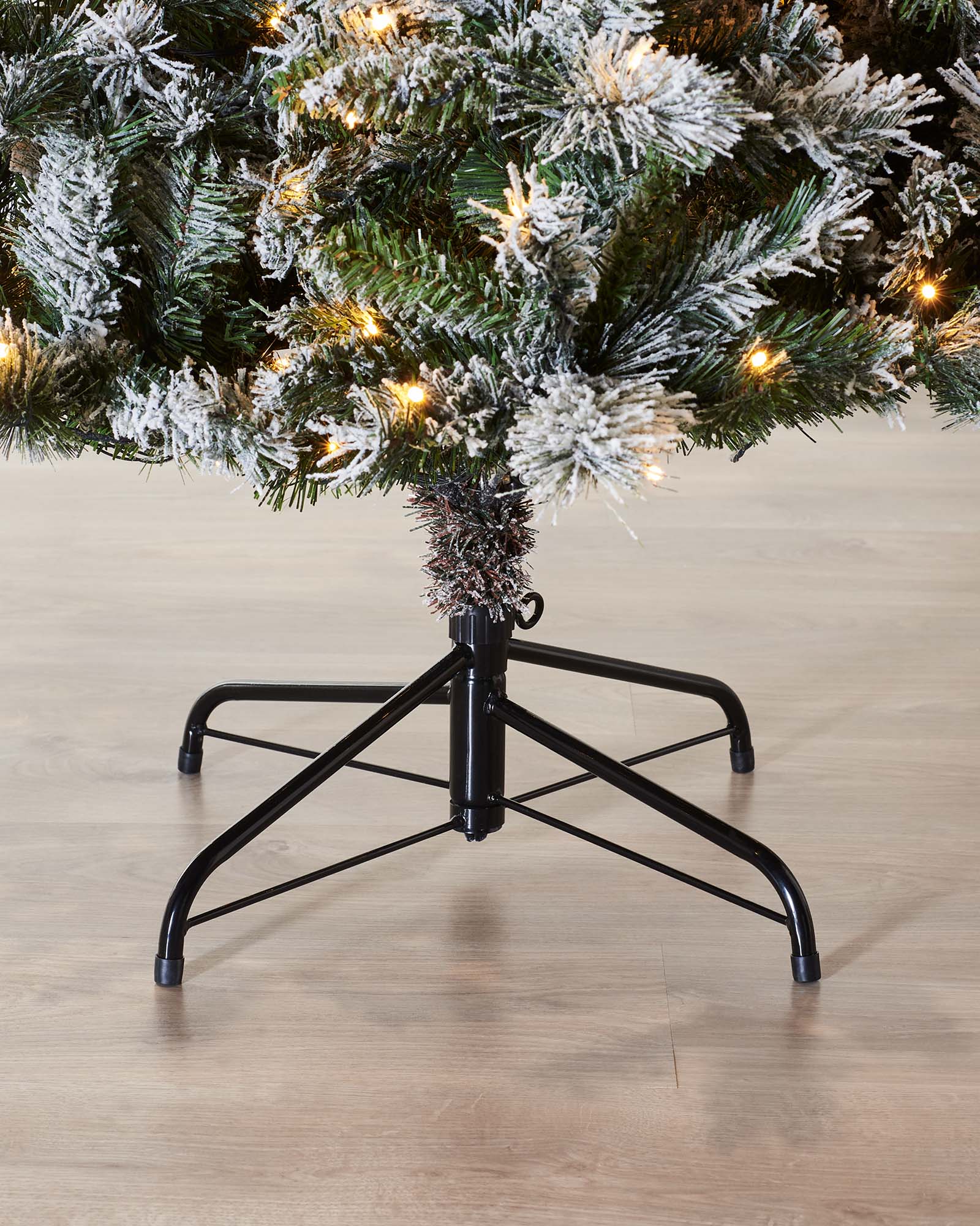 Close-up of the black metal tree stand beneath a pre-lit snow flocked spruce Christmas tree.
