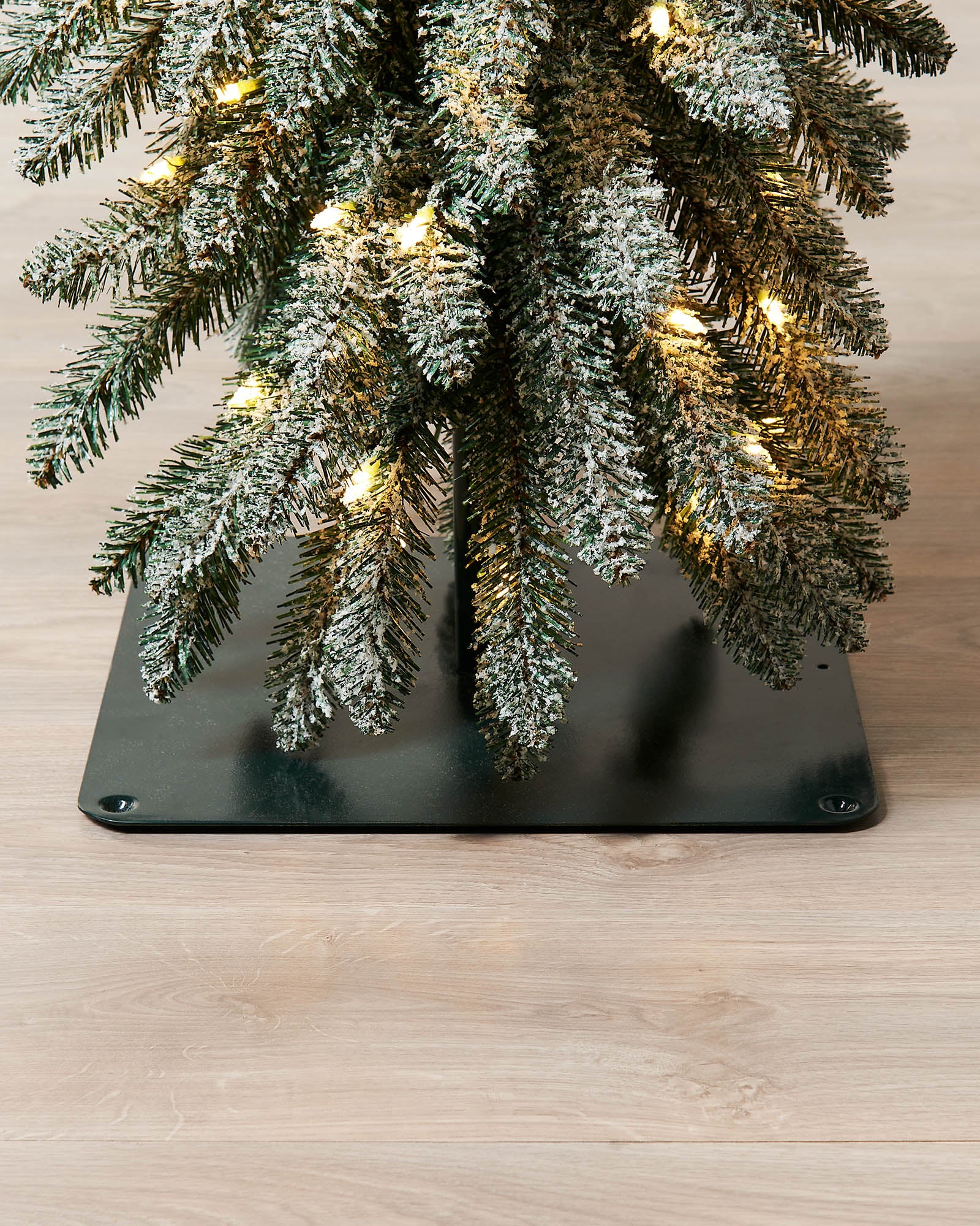 Base close-up of the snow-flocked alpine Christmas tree showing branches, lights and square metal base.