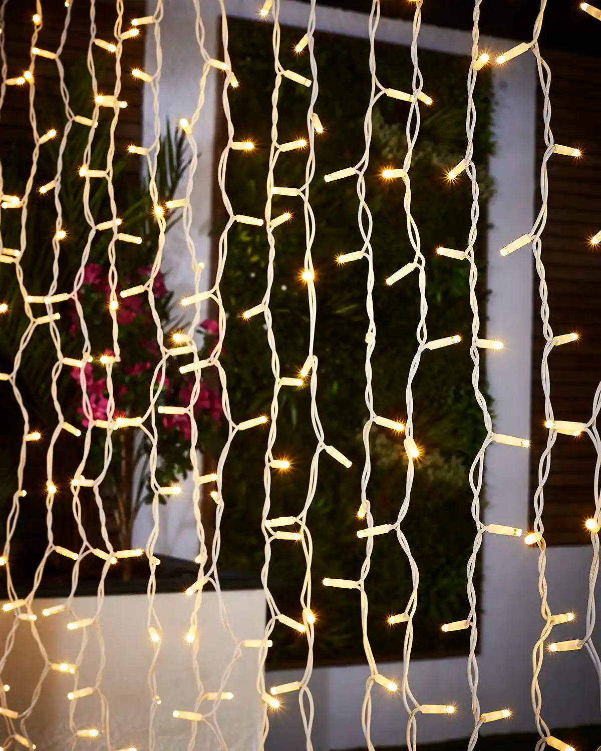 Warm white curtain light strands on white cable hanging in front of outdoor greenery.