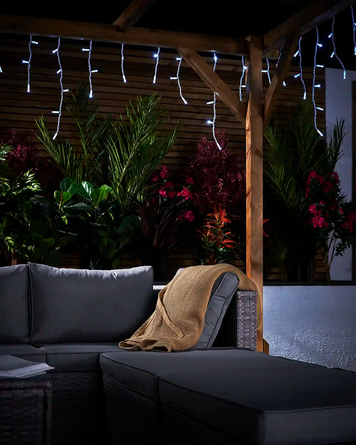 White icicle lights hanging from a pergola above garden seating and plants.