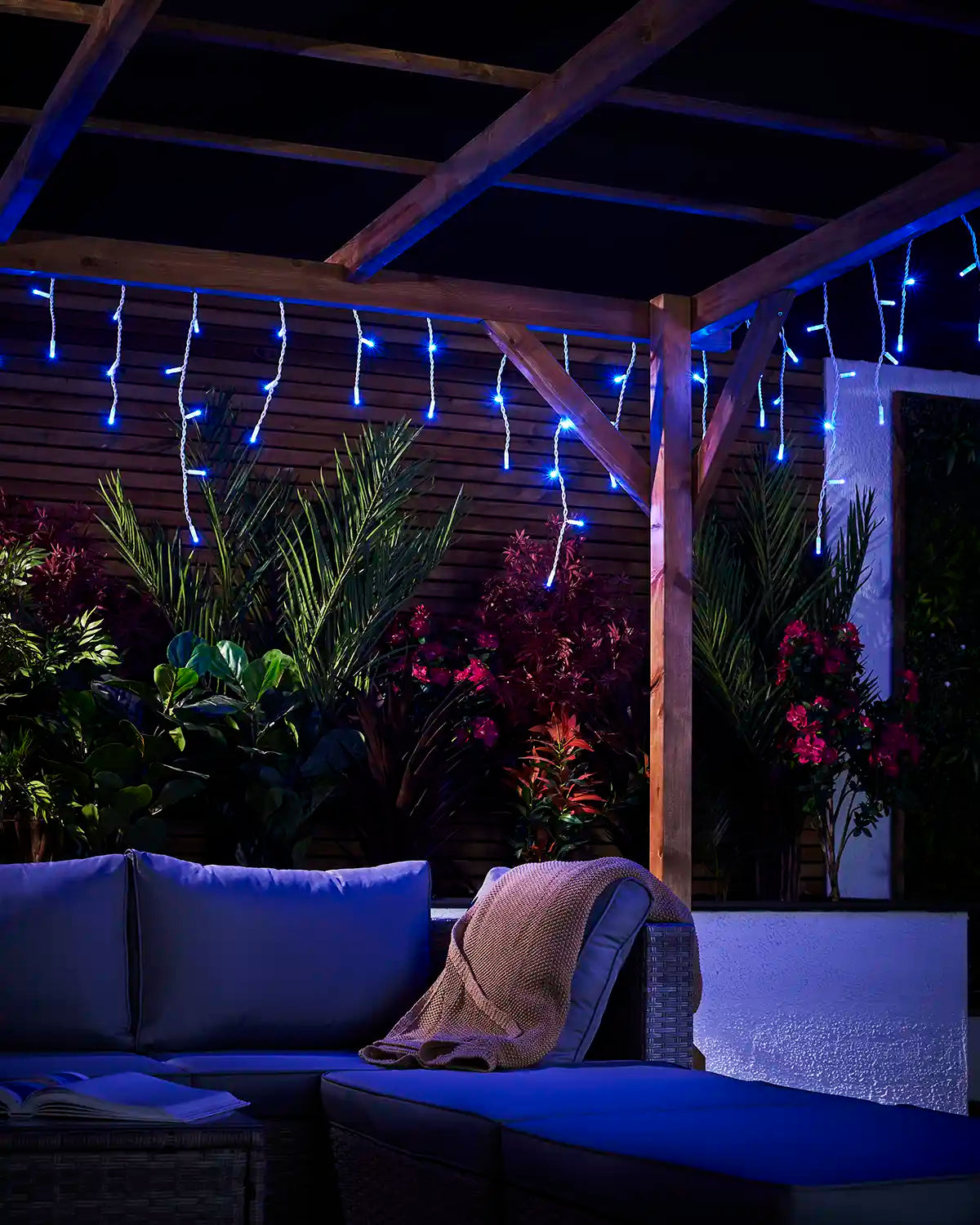 Blue icicle lights hanging from a wooden pergola above garden plants and seating.