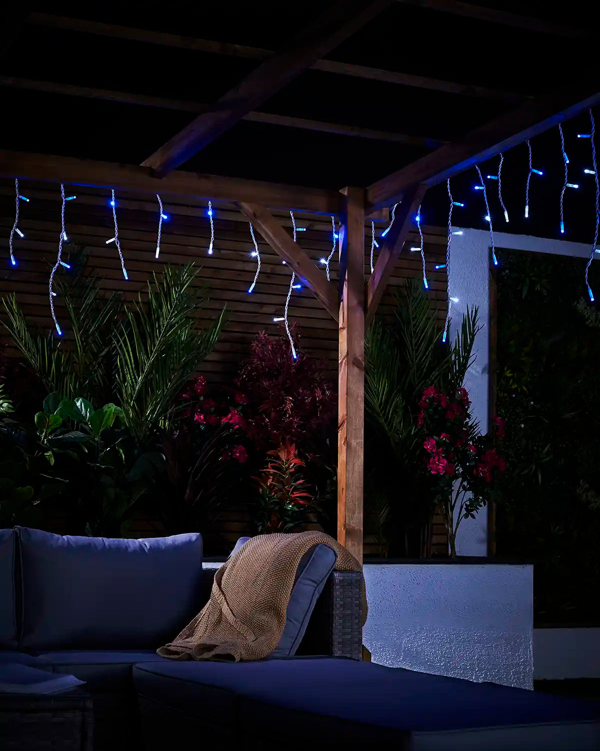 Blue and white icicle lights hanging from a wooden pergola in a garden seating area.
