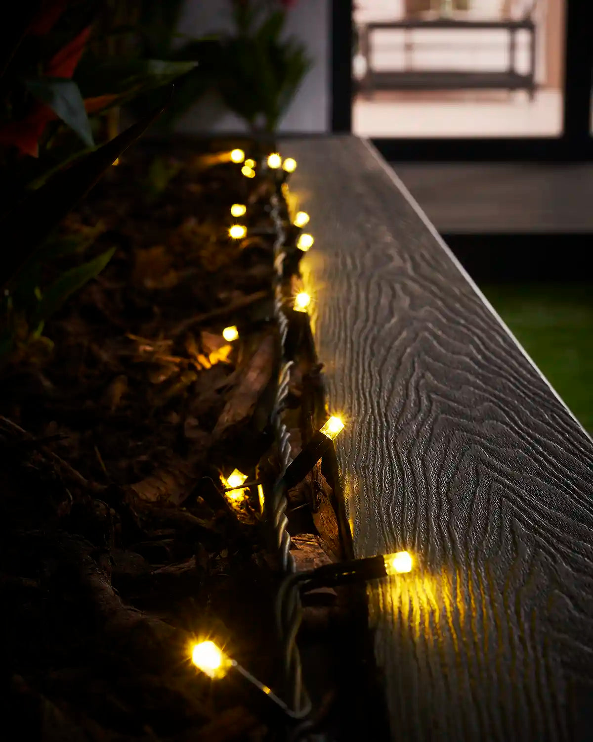 Gold LED string lights on black cable running along an outdoor planter rail.
